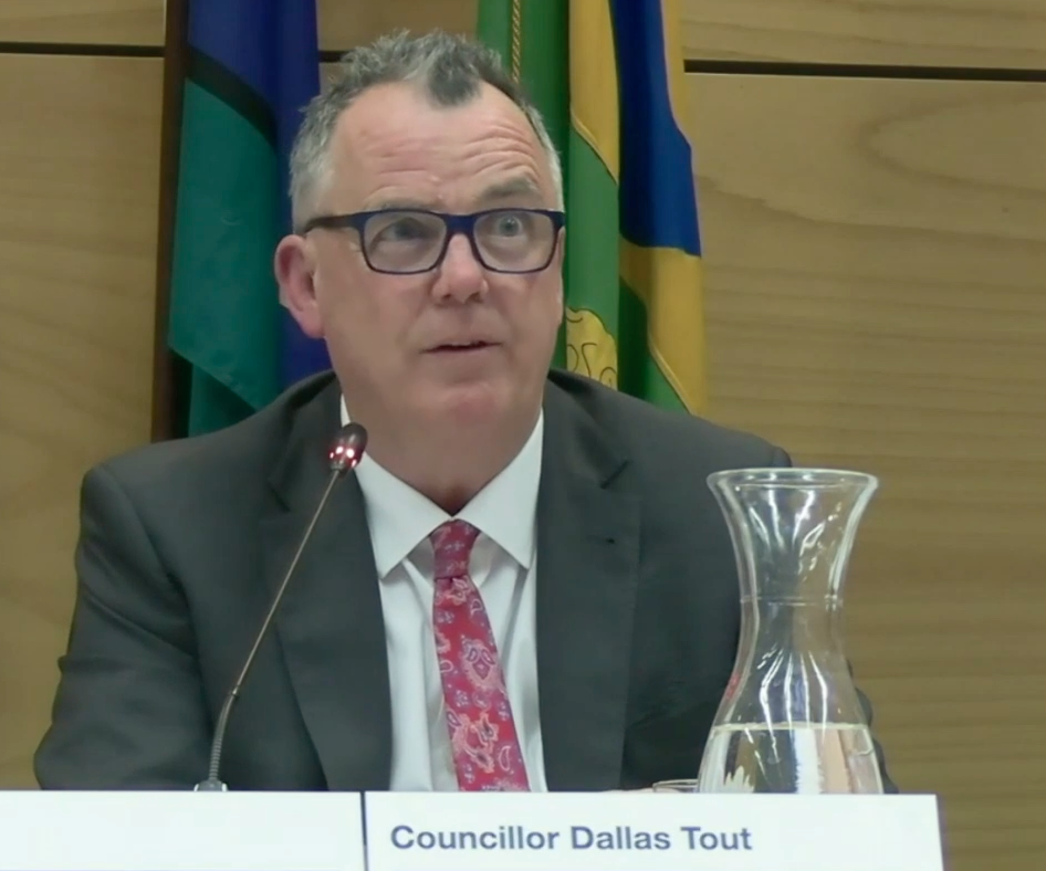 man in a suit sitting at a table in a council chamber